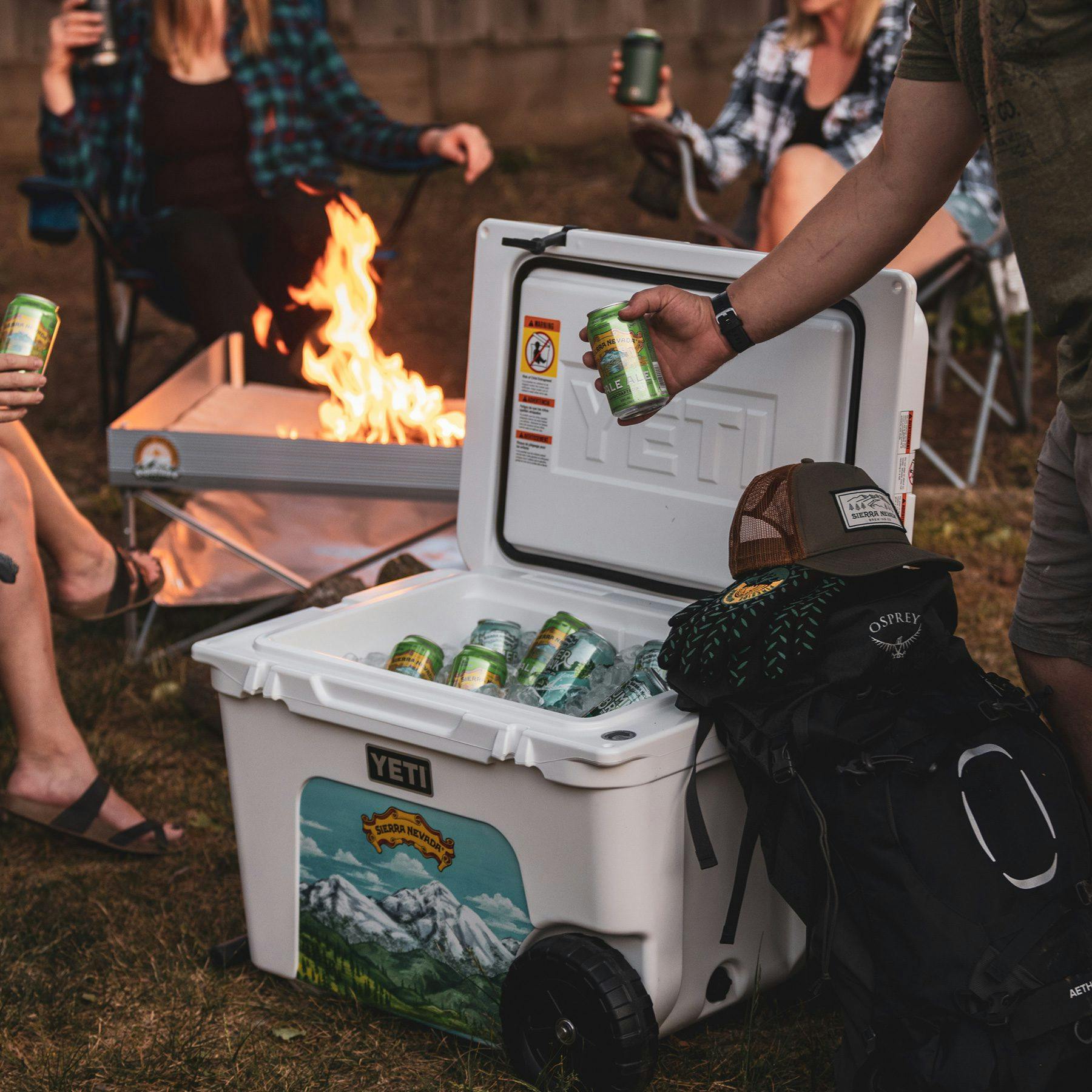 Person pulling a can of Sierra Nevada Pale Ale out of a YETI cooler that's in front of a fire pit
