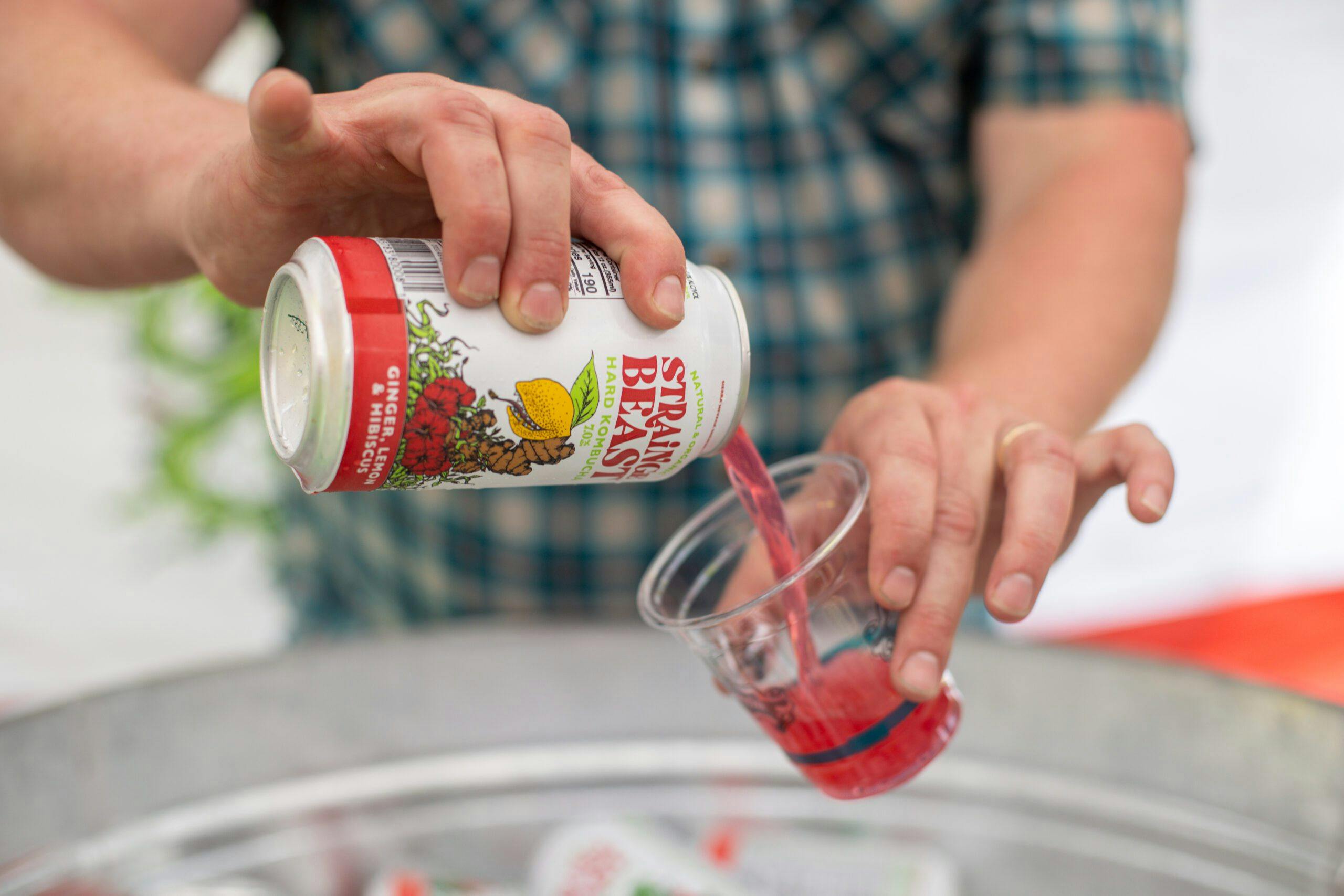 A person pours a can of Strainge Beast Ginger, Lemon and Hibiscus kombucha into a glass