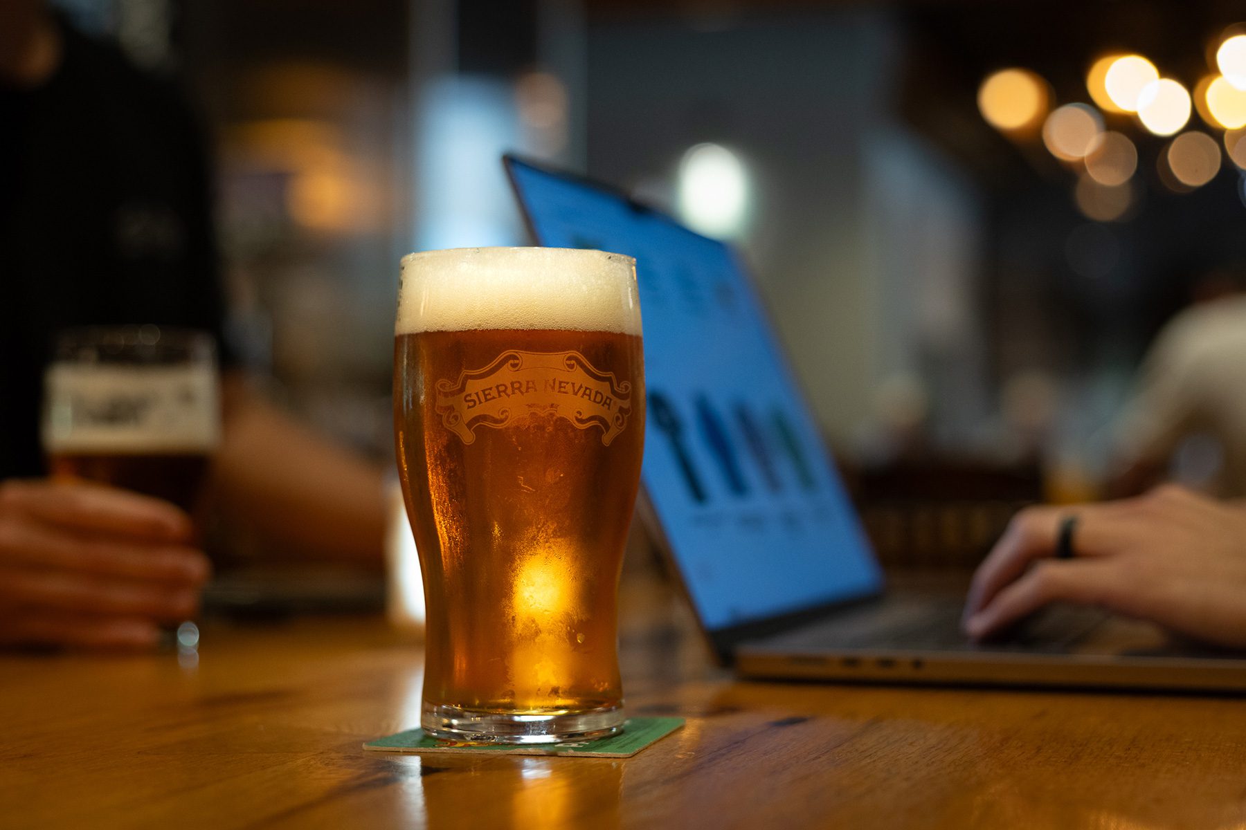 A full glass of beer in front of a person typing on a laptop