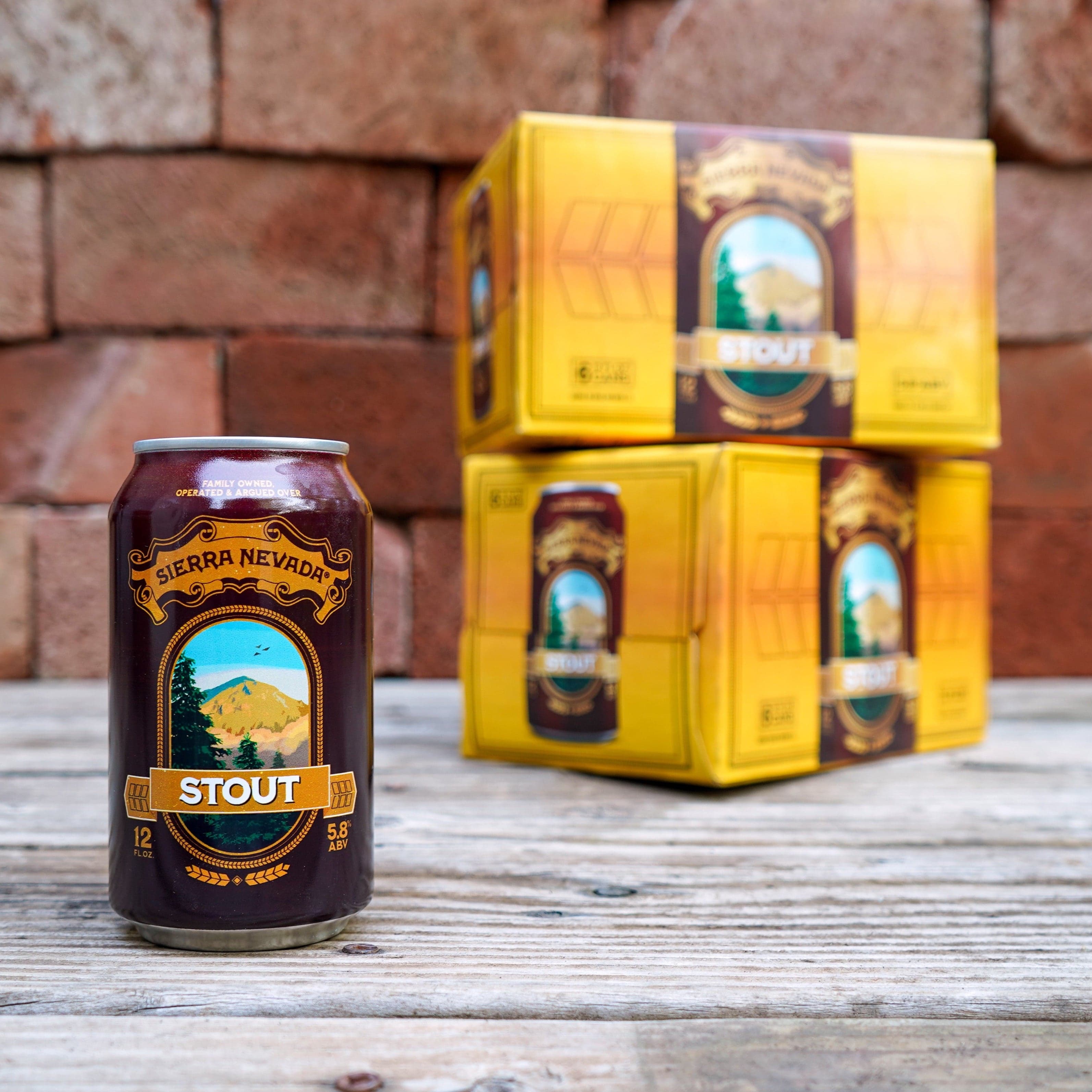 A 12oz. can of Sierra Nevada Stout in the foreground on a wooden tabletop with two 6-pack cases stacked behind it in the background