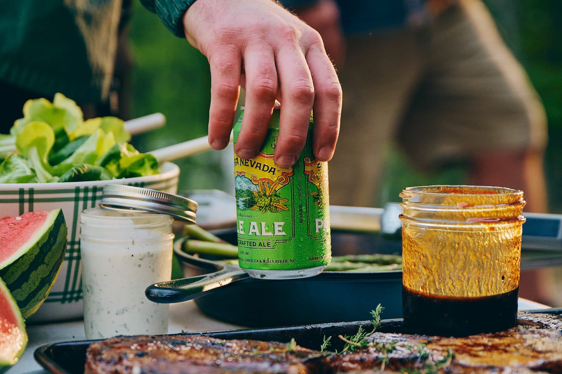 A hand picking up a can of Sierra Nevada Pale Ale from an outdoor table with food