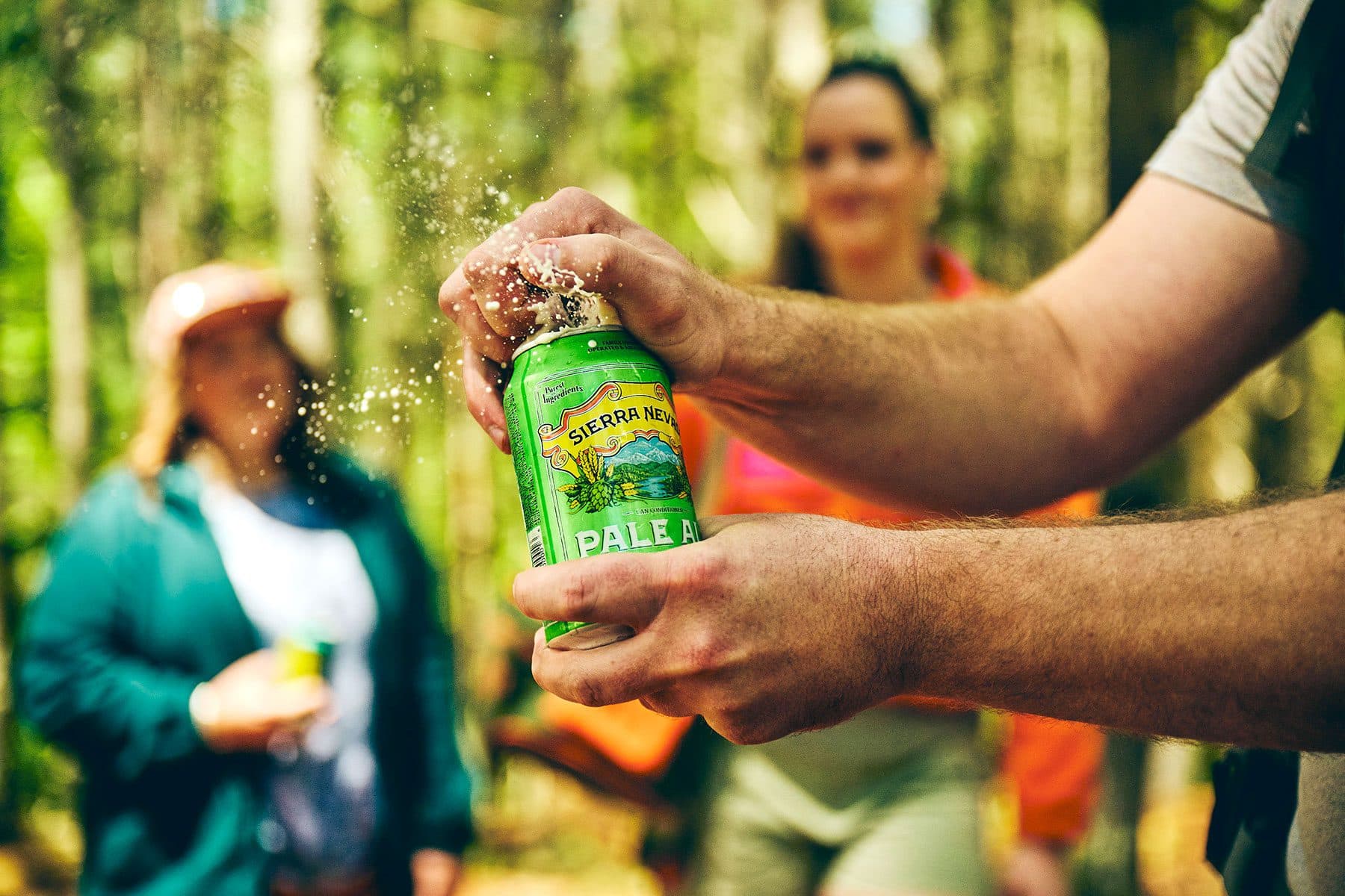 Hands opening a can of Sierra Nevada Pale Ale with two friends in the background surrounded by forest trees