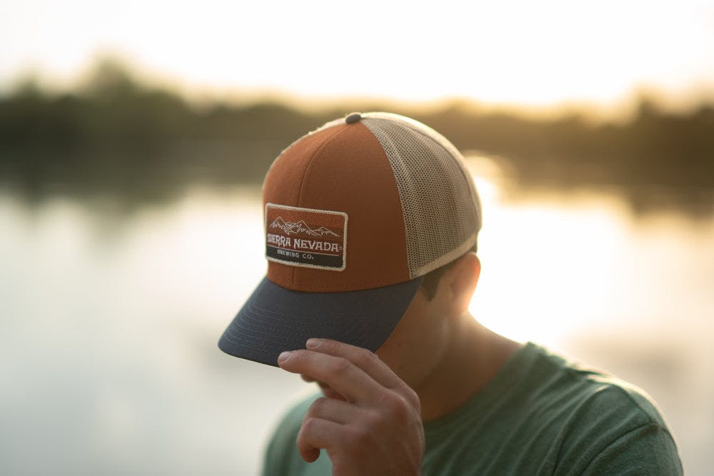 A man wears the Sierra Nevada Rusty Mountain Gradient Trucker Hat while walking along the banks of a river.