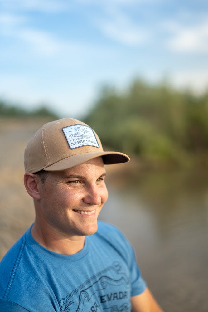 A man wears the Sierra Nevada Line Art Hat on an outdoor adventure to the edge of a river.
