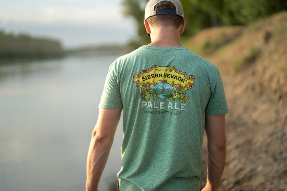 A man walks along a peaceful river wearing the Sierra Nevada Brewing Co. Pale Ale t-shirt, as viewed from behind.