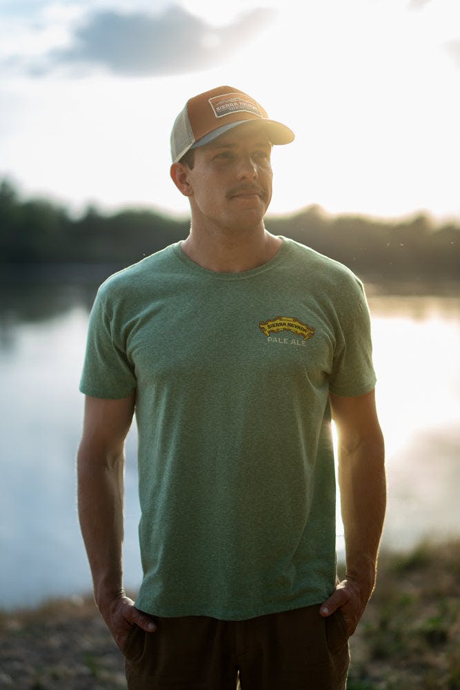 A man wears the Sierra Nevada Pale Ale T-Shirt while standing alongside a river.