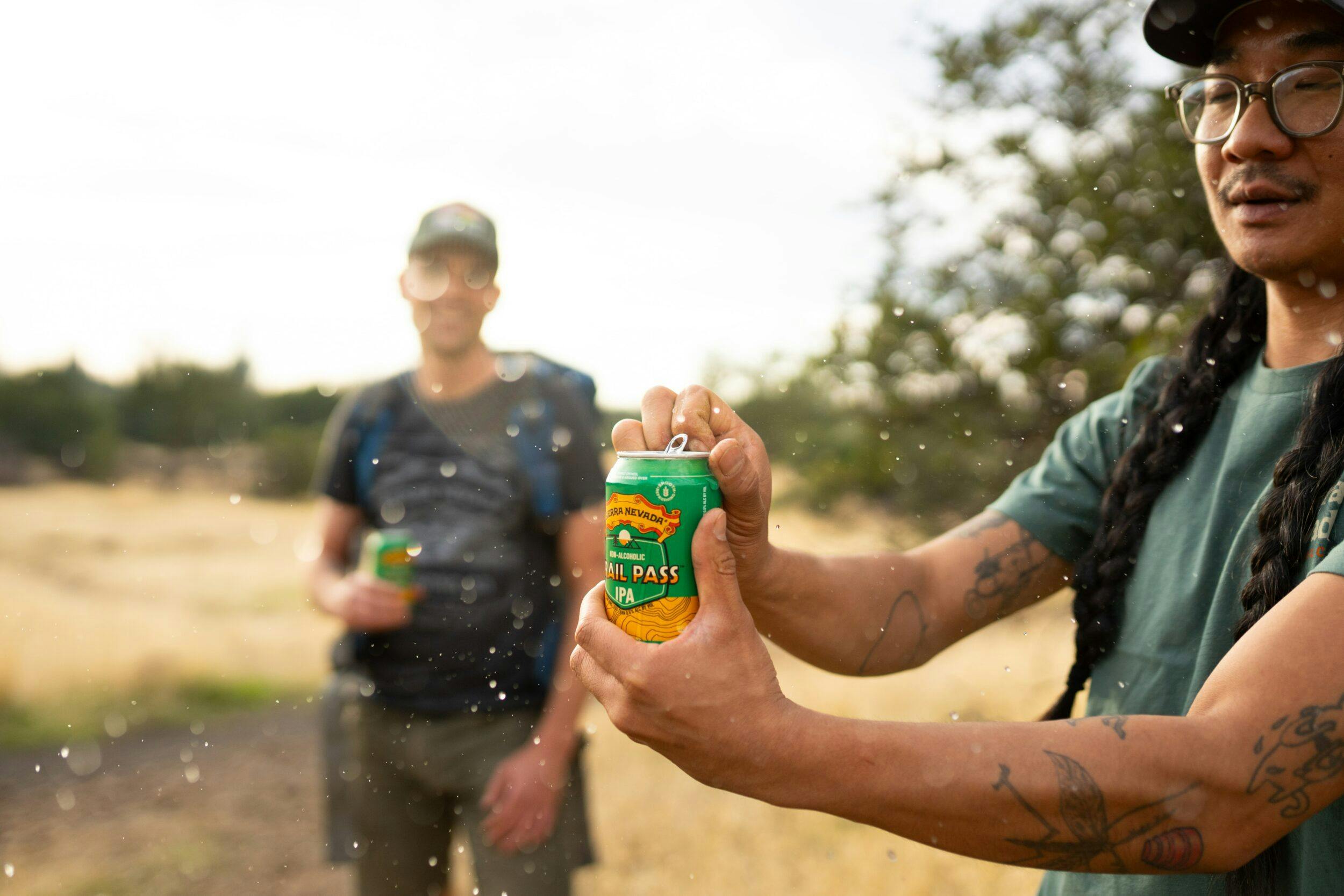Two male hikers on a trail opening a can of non-alcoholic Sierra Nevada Trail Pass IPA