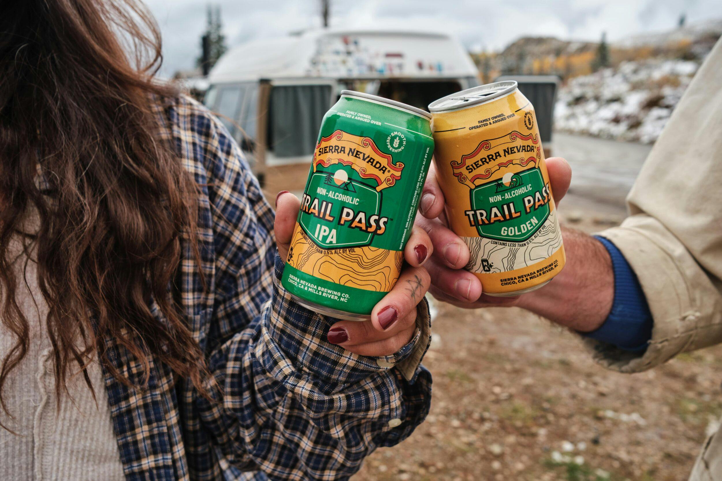 A young woman and a young man, outside of a camper van, toasting cans of non-alcoholic Sierra Nevada Trail Pass IPA and Trail Pass Golden